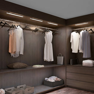 A modern walk-in closet with dark wood shelves, white and beige clothing, baskets, towels, a pillow, a shopping bag, and soft overhead lighting from the Artika Outline under cabinet LED light 3CCT.
