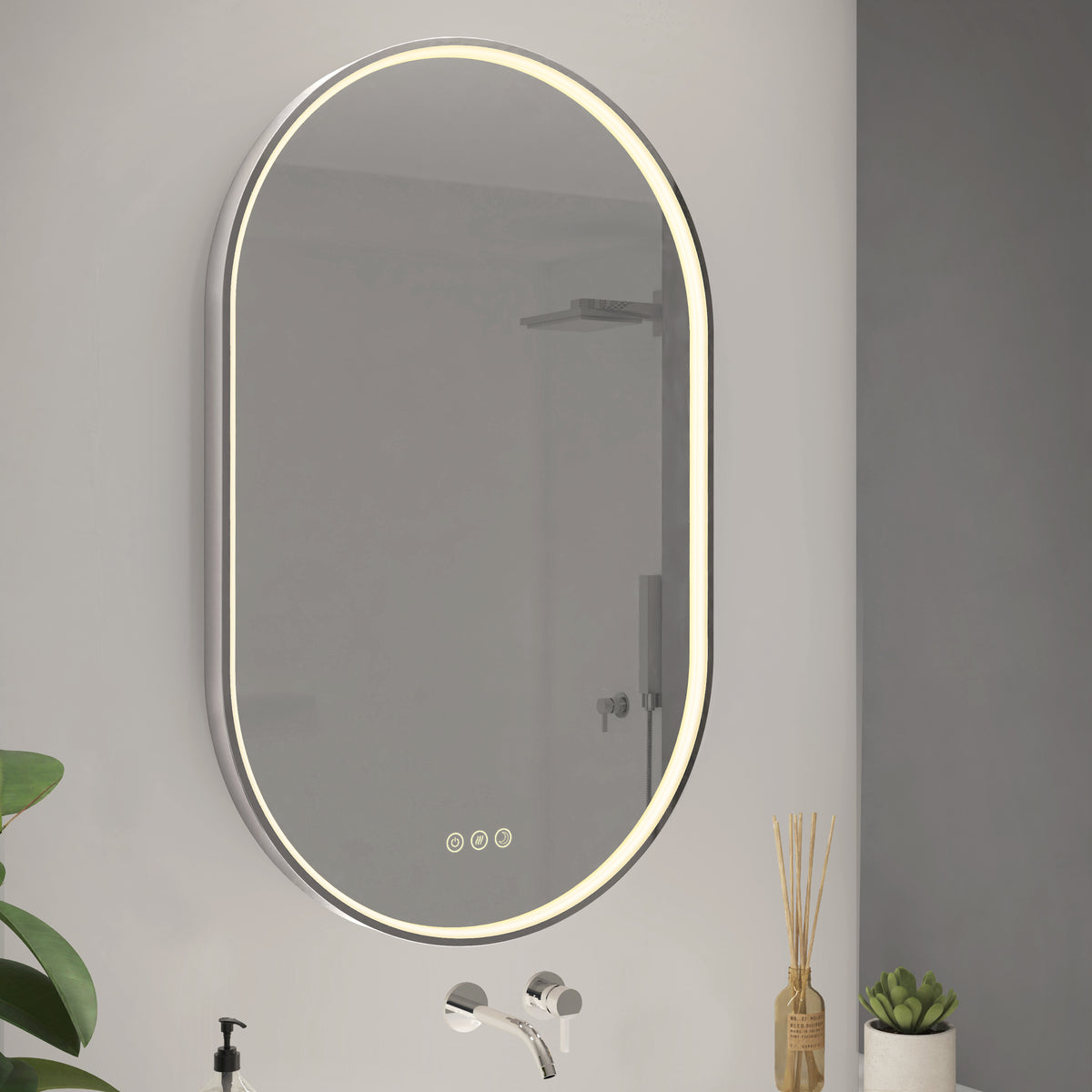 An oval-shaped, wall-mounted bathroom mirror with an illuminated LED border. Below the mirror are touch controls. A faucet, green plant, reed diffuser, and small potted succulent sit on the nearby counter.