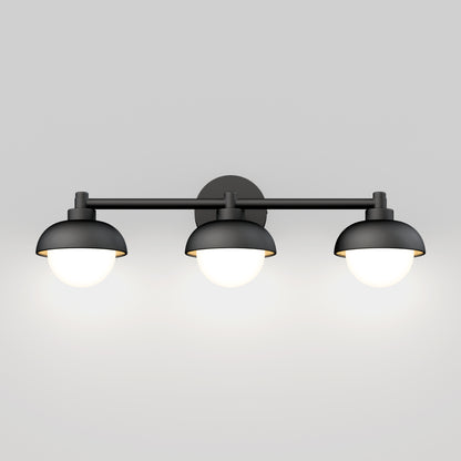 The Artika Boden LED mid-century bathroom vanity light features three dome-shaped lights on a horizontal bar, finished in modern black, softly illuminating a plain light gray wall.