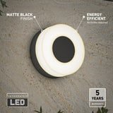 Modern round wall light with a matte black finish mounted on a textured wall. It is glowing softly, labeled energy efficient and integrated LED. Icons highlight 5 years warranty and no bulbs required. Some green plants are visible below.