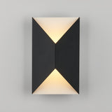 A modern wall sconce with a geometric black design emits soft, warm light from the top and bottom against a plain gray background.
