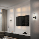 Modern living room with a wall-mounted flat-screen TV, two cylindrical wall sconces, a gray sofa, floor-to-ceiling curtains, and a minimalist cabinet with decorative vases and branches.