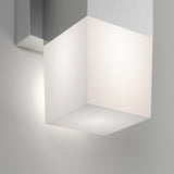 A modern wall-mounted light fixture with a frosted glass cube shade and a metallic base, softly illuminating the surrounding white wall.