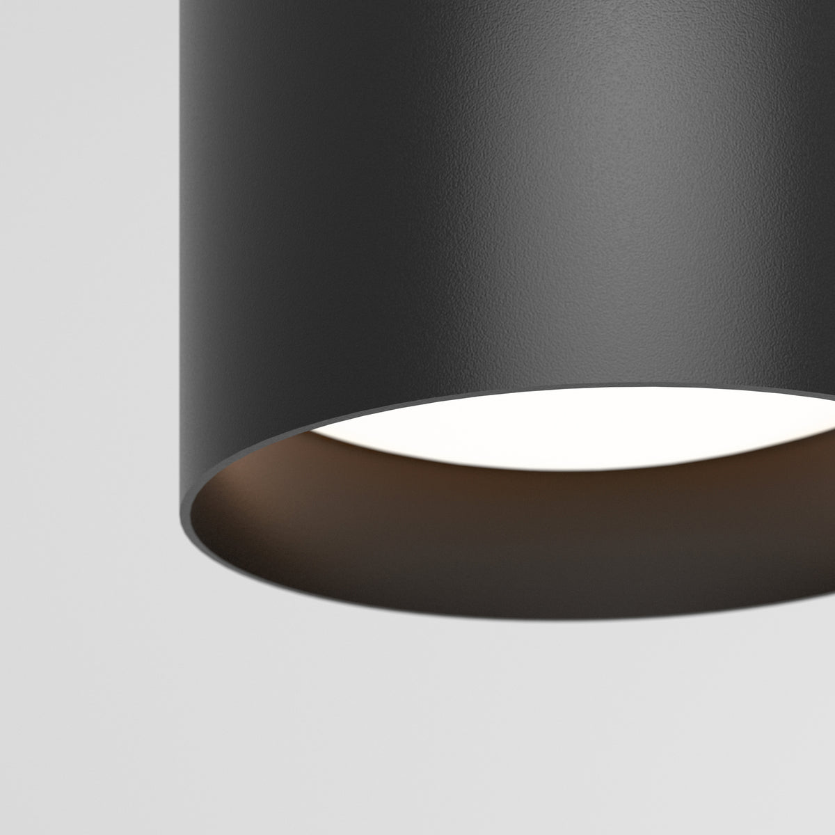 Close-up of a modern, cylindrical black ceiling light fixture with a smooth texture, hanging against a plain white background. The light is turned on, illuminating the inside of the fixture.