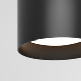 Close-up of a modern, cylindrical black ceiling light fixture with a smooth texture, hanging against a plain white background. The light is turned on, illuminating the inside of the fixture.