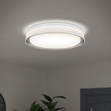 A modern, round ceiling light fixture emits a soft, bright glow in a minimalist room with white walls and a glimpse of green plant leaves below.