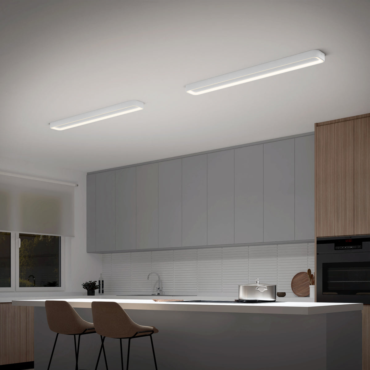 A modern kitchen with light gray cabinets, wood accents, two beige bar stools at a white island, and two rectangular ceiling lights. A window with a white blind lets in natural light.