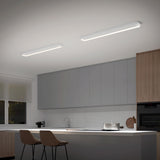 A modern kitchen with light gray cabinets, wood accents, two beige bar stools at a white island, and two rectangular ceiling lights. A window with a white blind lets in natural light.