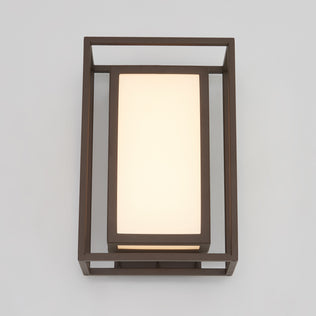 The Artika Ghost indoor-outdoor LED wall light features a rectangular frosted glass shade framed by a dark metal geometric structure, stylishly mounted on a light gray wall.