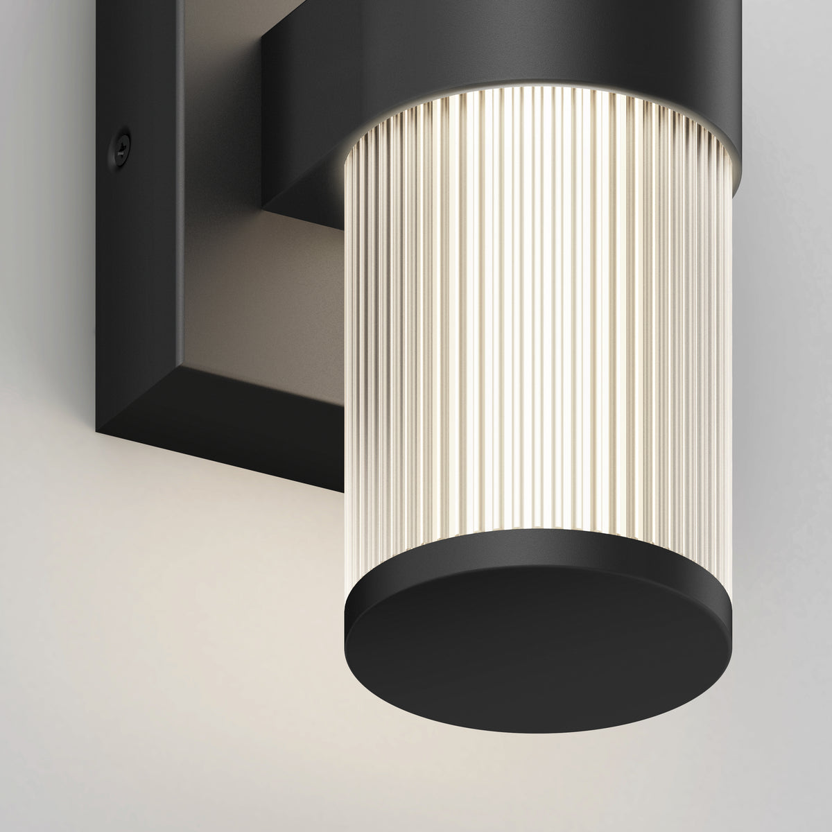 A close-up view of a modern wall-mounted light fixture with a black metal base and a cylindrical, ribbed glass shade emitting a soft, warm glow.