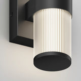 A close-up view of a modern wall-mounted light fixture with a black metal base and a cylindrical, ribbed glass shade emitting a soft, warm glow.