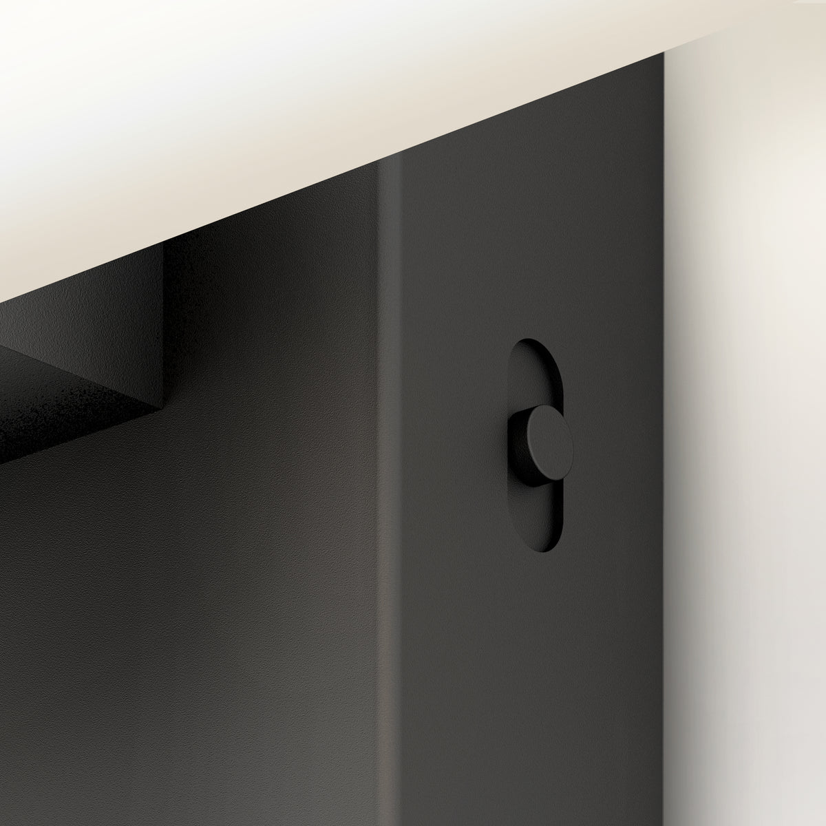 Close-up of a black metal object with a circular button or knob on its surface, partially obscured by a light-colored flat panel above it. The design appears minimalistic and modern.