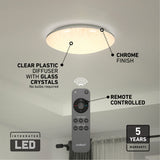 Round LED ceiling light with a chrome finish and clear plastic diffuser containing glass crystals, shown illuminated on a ceiling. A remote control is displayed in the foreground. Warranty information is also visible.