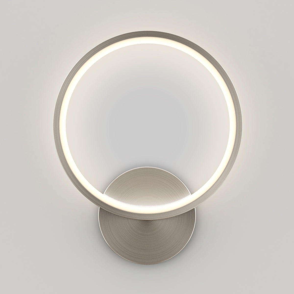 The Artika Solis modern LED indoor-outdoor wall light 5 CCT features a circular ring on a metallic base, emitting a soft white glow—perfect for accent lighting in contemporary spaces with light-colored walls.