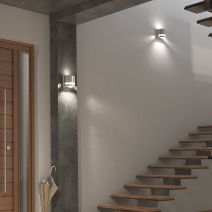 Modern interior with a wooden staircase, a tall grey concrete pillar, two Artika Valor Round LED indoor-outdoor wall lights, a wooden door with metal accents, and an umbrella stand by the entrance. The walls and decor are minimalistic.