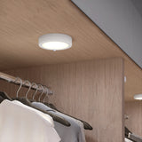 A modern, circular LED ceiling light is installed on a wooden surface above a clothes rack holding white and dark shirts in a closet. Another similar light is visible further back.