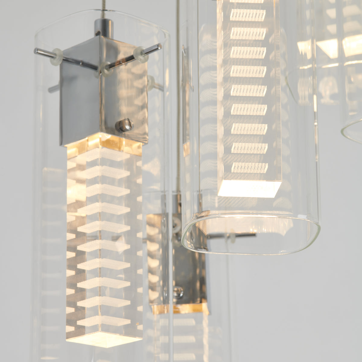A close-up of a modern pendant light fixture with a square, layered design inside a clear cylindrical glass shade, emitting a soft, warm glow. The fixture has metal accents and a minimalist aesthetic.
