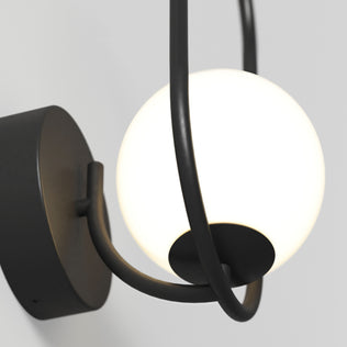 Close-up of the Artika Bloom LED Sconce 3 CCT, featuring a black metal fixture and a round white frosted glass globe that emits soft light, mounted on a light gray wall.