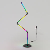 The Artika Fitch smart foldable RGB LED gaming floor lamp, featuring a modern zigzag shape and black base, displays multiple colors while plugged into a wall outlet against a plain light-gray background.