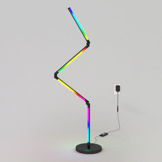 The Artika Fitch smart foldable RGB LED gaming floor lamp, featuring a modern zigzag shape and black base, displays multiple colors while plugged into a wall outlet against a plain light-gray background.