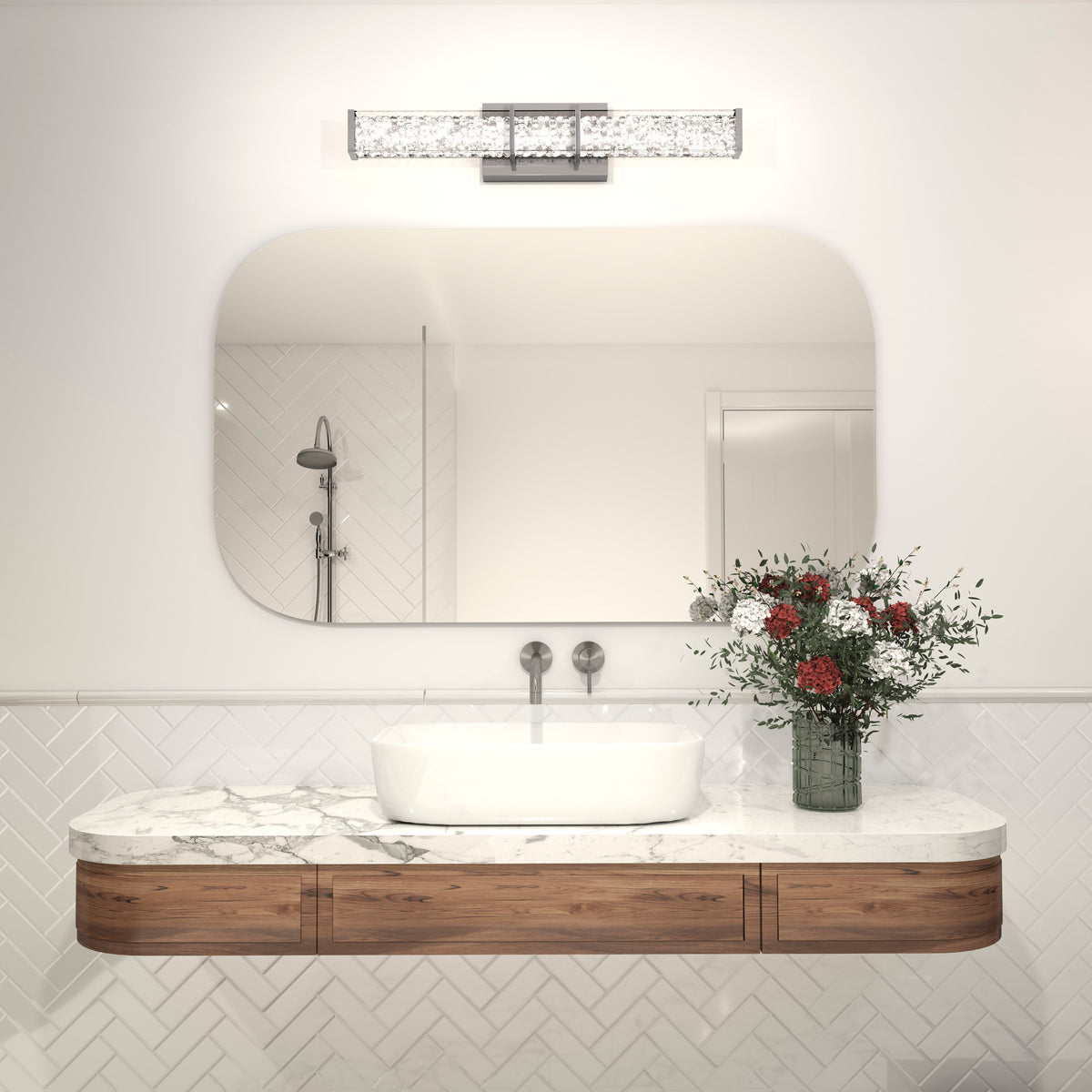 Modern bathroom with a marble countertop, wooden cabinet, rectangular sink, wall-mounted faucet, a vase of red and white flowers, large mirror, and contemporary light fixture above. White tiles in a herringbone pattern on the wall.