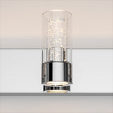 A modern wall-mounted light fixture with a cylindrical glass shade and chrome base, emitting warm white light with a sparkling effect inside the glass.