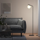 A modern living room with a gray sofa, a light gray pillow, a black floor lamp with a round white shade, a plant, a glass of water on a round table, and abstract wall art. Soft lighting creates a cozy atmosphere.