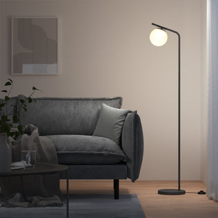 A modern living room features a gray sofa, light gray pillow, geometric wall art, coffee table with a glass, a plant, and the Artika Clayton dimmable LED modern mid-century floor lamp with a glowing round bulb. Light filters through sheer curtains.