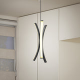 A modern pendant light with two curved, intersecting black and white LED bars hangs from the ceiling in a minimalist room with beige walls, cabinetry, and a window with layered blinds.