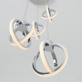Modern pendant light fixture with four spherical, chrome rings intertwined and illuminated, suspended from the ceiling by thin cables against a light background.