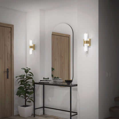 A modern entryway featuring a black console table, tall oval mirror, potted plant, stacked books, bowl, and two Artika Dorado LED contemporary wall sconces with gold and glass accents beside a wooden door.