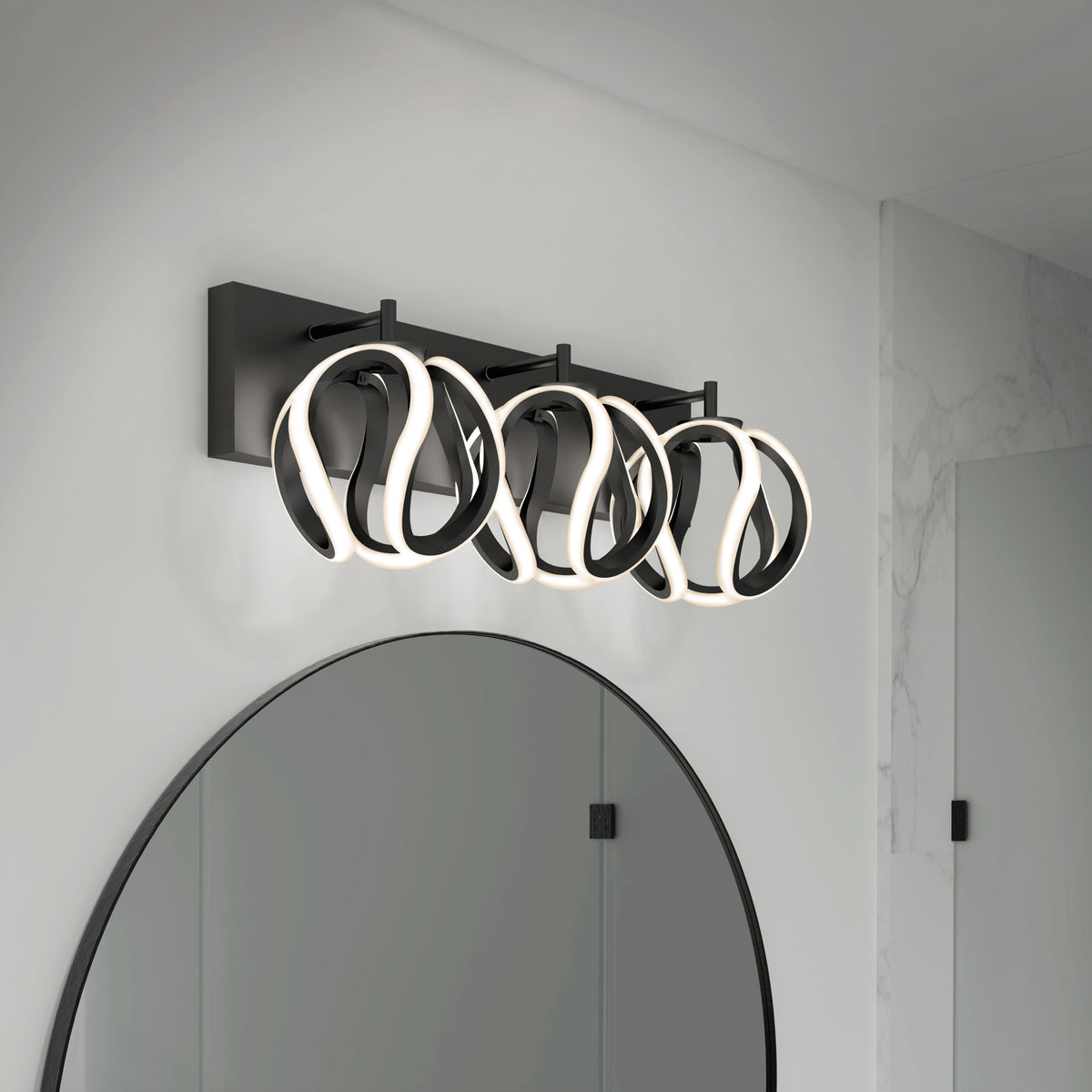 A modern bathroom light fixture with three abstract, looped LED lights mounted above a round mirror on a white wall with marble accents.