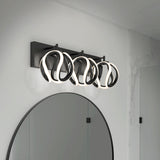 A modern bathroom light fixture with three abstract, looped LED lights mounted above a round mirror on a white wall with marble accents.