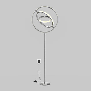 The Artika Pivot modern LED floor lamp 3 CCT features a sleek silver finish, circular elements, and an illuminated ring, shown plugged into a wall socket against a plain gray background.