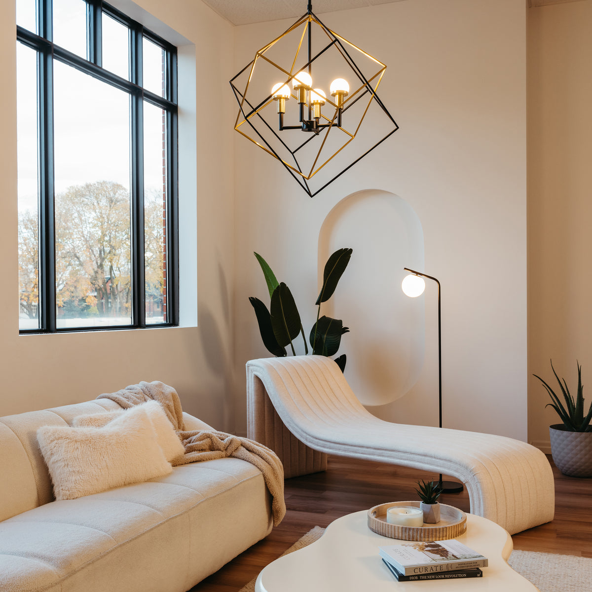 A modern, cozy living room with a cream sofa, a sculptural chaise lounge, potted plants, a geometric gold chandelier, a floor lamp, large windows, and a white coffee table with a tray and book.
