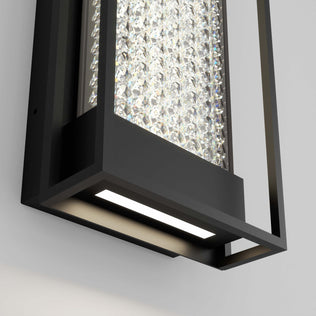 A close-up of the Artika Glam indoor-outdoor LED wall light features a black metal frame, clear crystal accents, and a bright LED shining downward against a white wall.