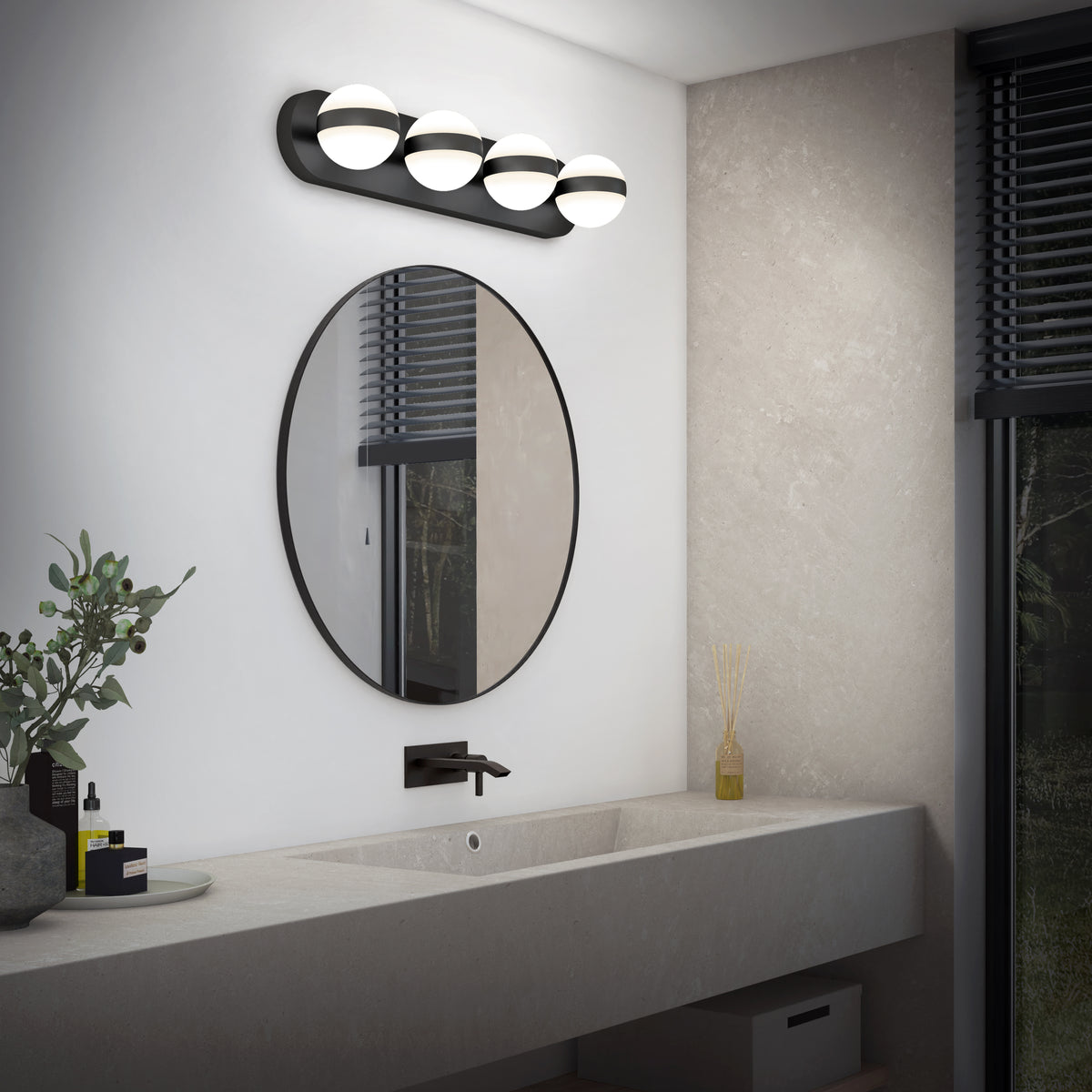 A modern bathroom with a round mirror above a long, light gray sink, matte black faucet, and four-globe wall light. There are toiletries, a small plant, and reed diffuser on the countertop. A window with blinds is on the right.