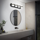 A modern bathroom with a round mirror above a long, light gray sink, matte black faucet, and four-globe wall light. There are toiletries, a small plant, and reed diffuser on the countertop. A window with blinds is on the right.