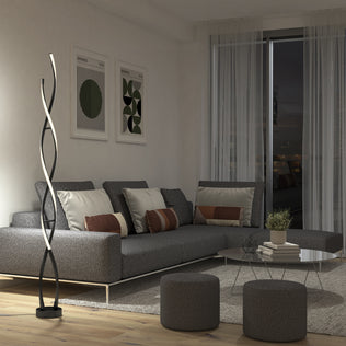 A modern living room with a gray sectional sofa, decorative pillows, two round ottomans, abstract wall art, a round coffee table, sheer curtains, a large window, and the Artika Swirl modern LED floor lamp.