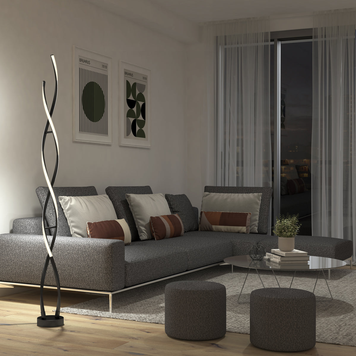 A modern living room with a grey sectional sofa, two round ottomans, geometric wall art, a tall twisted LED floor lamp, a round glass coffee table, and floor-to-ceiling windows with sheer curtains at night.