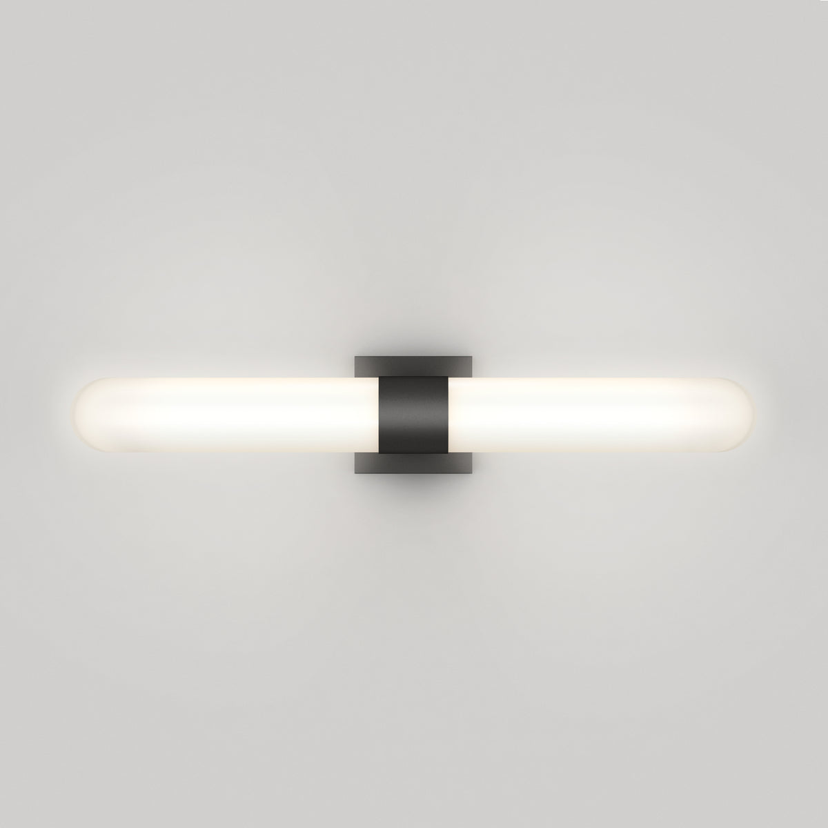 A modern wall-mounted light fixture with a horizontal, cylindrical white frosted glass shade and a central black rectangular bracket, illuminated against a plain white wall.