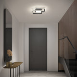 A modern hallway with a dark door, wooden side table holding a vase with dried flowers, large wall mirror, glass stair railing, wood panel accent wall, and geometric ceiling light fixture.