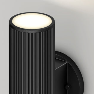 A close-up of the Artika Groove LED indoor-outdoor wall light, featuring a modern black ribbed cylindrical design and emitting soft white light from the top, mounted on a light-colored wall.