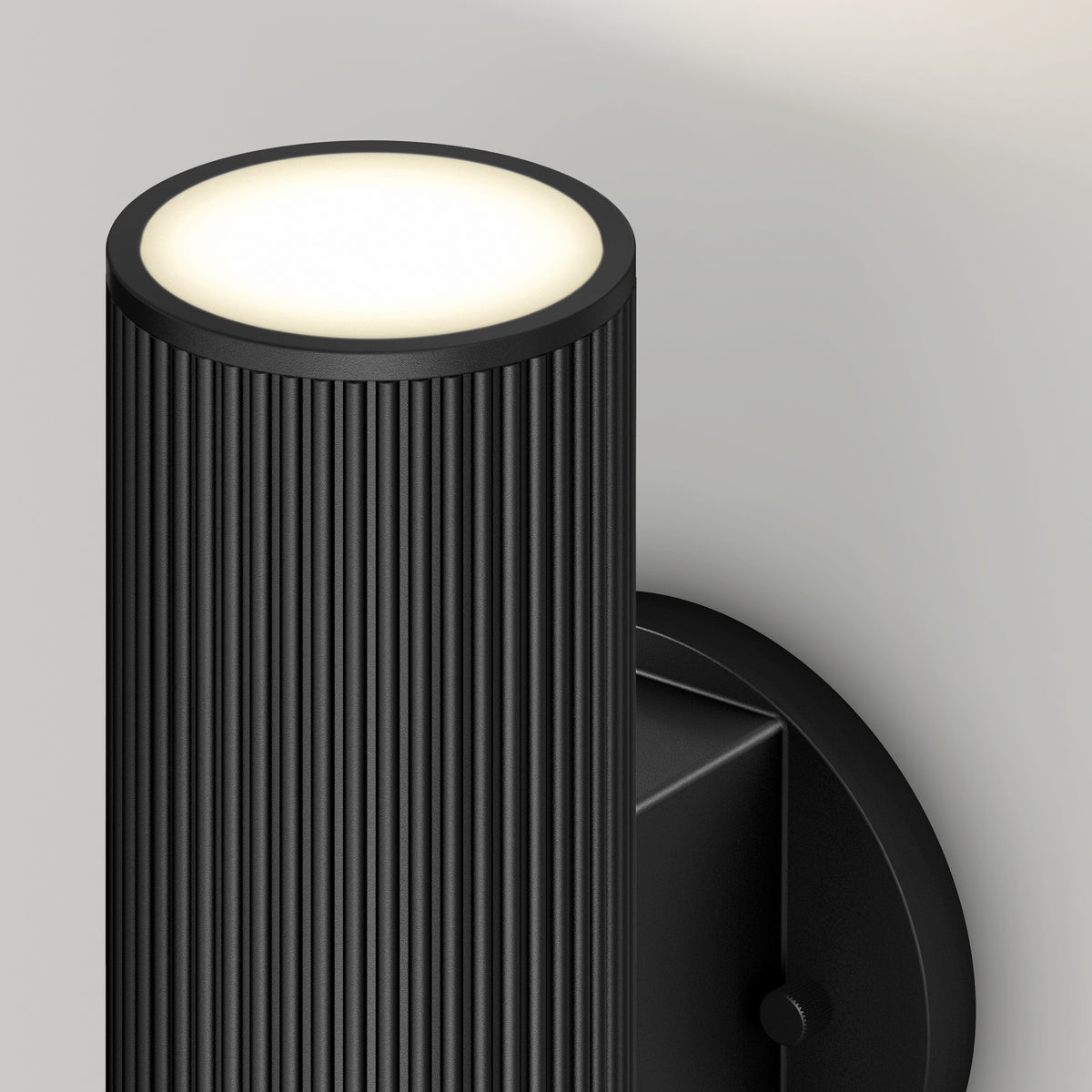 A modern black wall sconce with a ribbed cylindrical design emits soft white light from the top, mounted on a light gray wall.