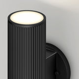 A modern black wall sconce with a ribbed cylindrical design emits soft white light from the top, mounted on a light gray wall.