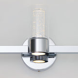 A modern wall sconce with a chrome base and cylindrical glass shade, featuring small bubbles inside the glass and an LED light glowing from the bottom.