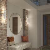 A modern entryway with exposed brick columns, a round mirror, wall sconces, a cushioned bench, two accent pillows, and a wooden door in the background. Warm lighting creates a cozy atmosphere.