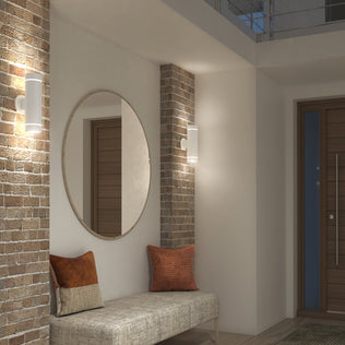 A modern entryway with exposed brick columns, a large round mirror, a cushioned bench with two accent pillows, an Artika C7 indoor-outdoor LED wall light 3 CCT, and a wooden front door with a vertical metal handle.