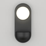 A modern wall sconce features a round white light at the top and a small, half-round black shelf below, all mounted on an oval dark gray backplate against a light gray wall.
