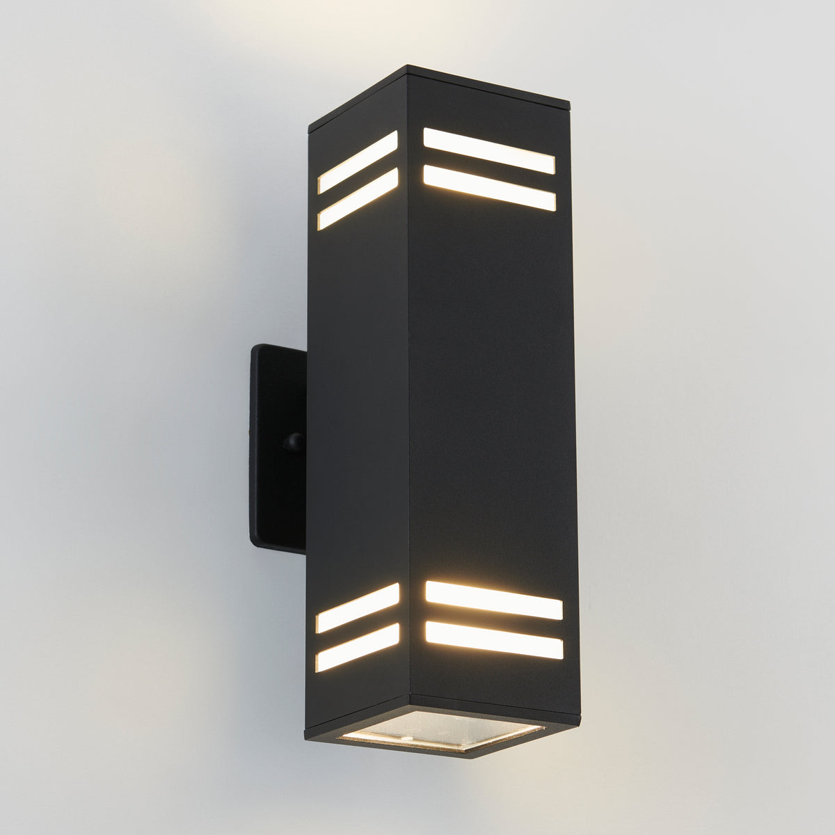 The Artika Strike indoor-outdoor wall light is a modern black rectangular sconce with horizontal slits near the top and bottom, mounted on a white wall and emitting soft white light from both ends and sides.